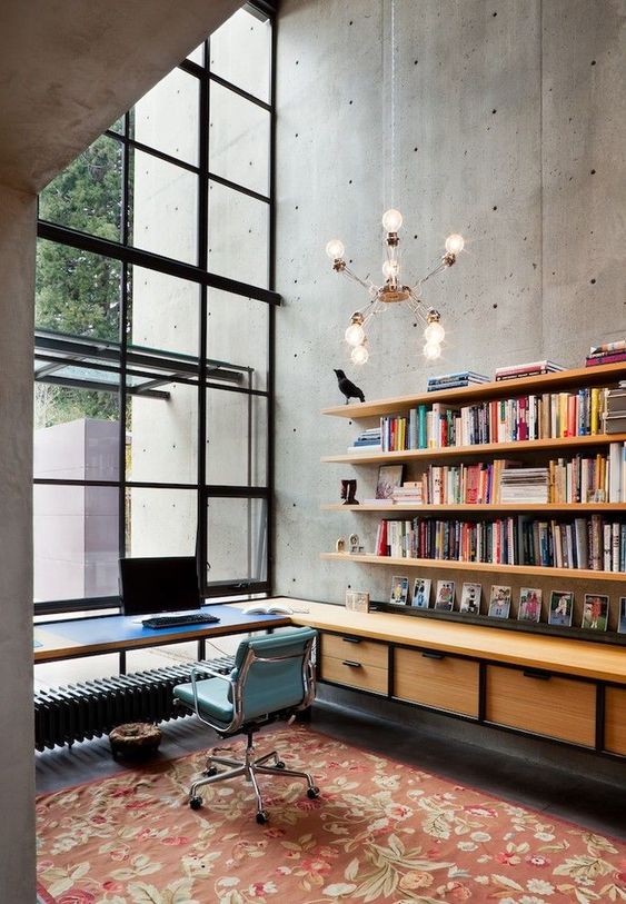 an industrial home office with concrete walls, a glazed wall, open shelves and a floating storage unit, a printed rug and a cool chandelier