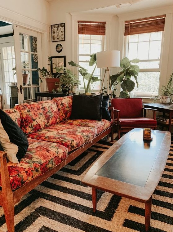 A mid century modern lviing room with a black and white rug, a floral sofa, rust colored chairs, potted plants and a floor lamp