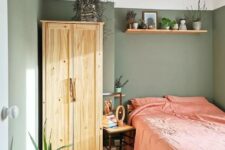 20 a small sage green bedroom with a stained wardrobe and a bed with coral bedding, open shelves, a chair and potted plants