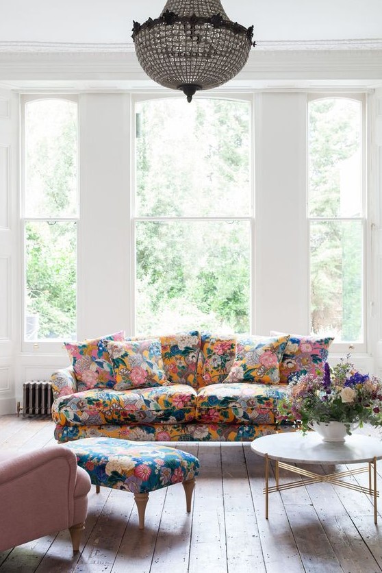 an airy living room with large windows, a floral sofa and a footrest, a pink chair, a coffee table and a crystal chandelier
