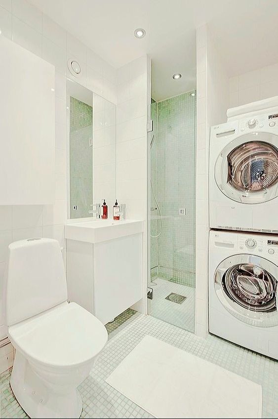 a minimal white bathroom clad with small tiles, with a stacked washing machine and dryer, a shower and a sleek vanity