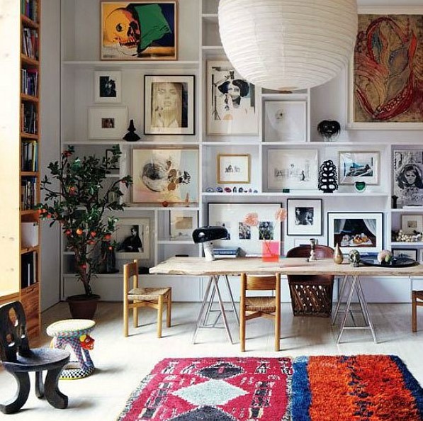 A double height space with a large gallery wall taking the whole wall, a bookcase, a living edge desk, several chairs and a bright rug