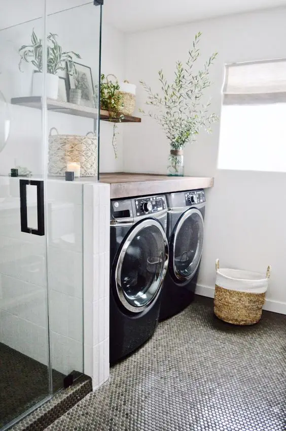 a modern bathroom with penny tiles, a glass enclosed shower, a washing machine and a dryer, greenery and a basket