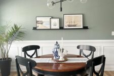 34 a stylish modern dining room with a sage green wall and paneling, a dark-stained table and chairs, a cool chandelier and some art