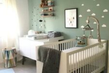 38 a cozy Nordic nursery with a sage green accent wall, white furniture, a pendant lamp and some shelves plus neutral bedding