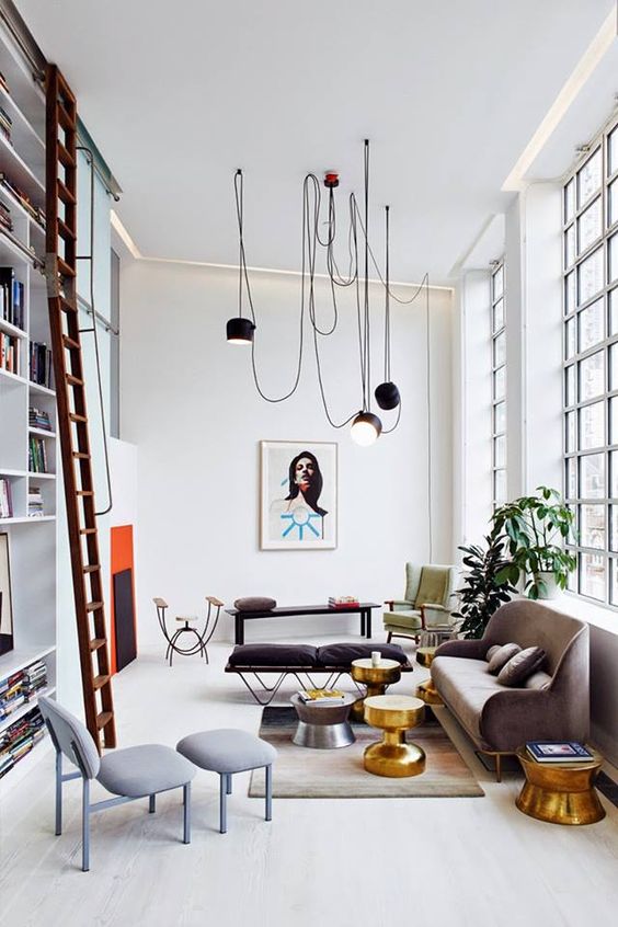a cool eclectic living room with a double-height bookcase and windows, a neutral sofa, a grey chair and a brown daybed, gilded tables and black lamps