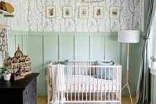 40 a lovely nursery with mint grene paneling and pretty wallpaper, a neutral crib and a black dresser, a chandelier and a pouf