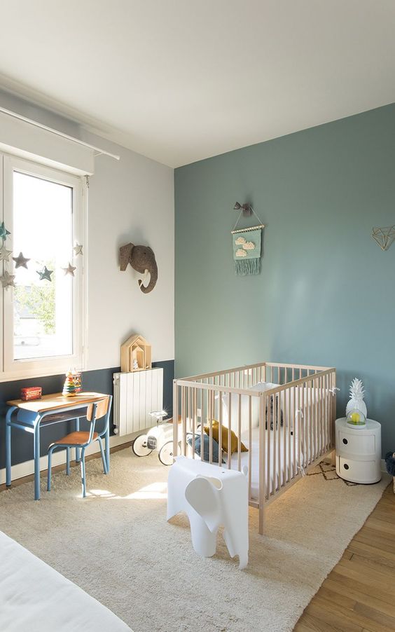 an airy Scandinavian nursery with a sage green accent wall, a wooden crib, a small desk and a chair, some lovely decor