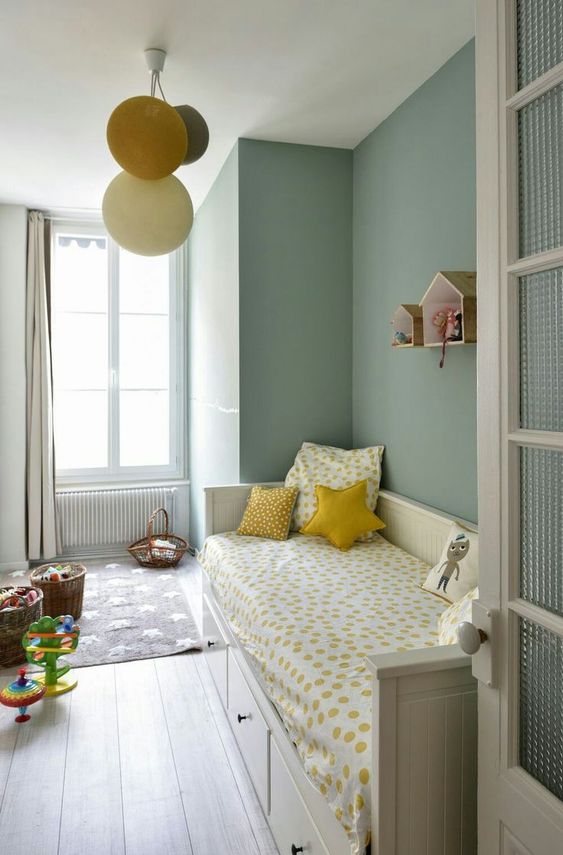 A chic sage green kids' room with a storage bed and bright bedding, baskets with toys and house shaped shelves