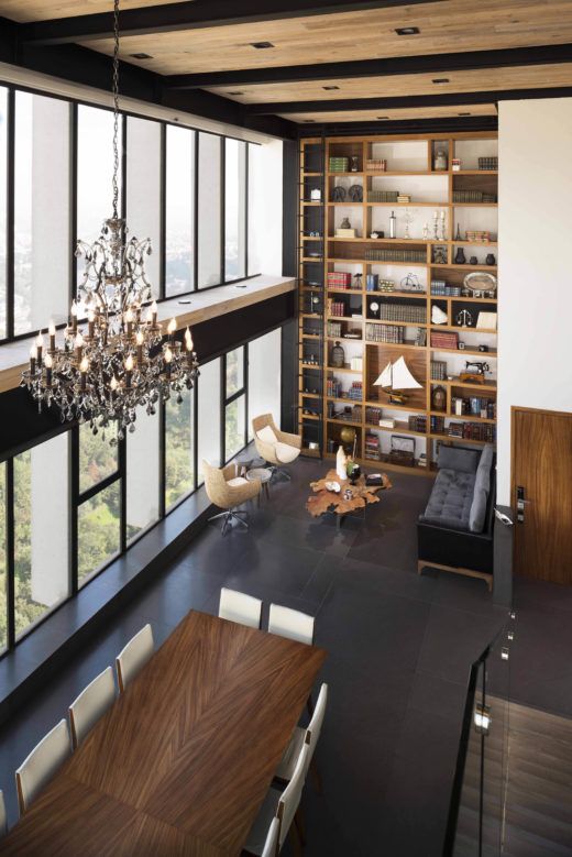 an open layout with a glazed wall, a large bookcase, a black sofa, neutral chairs, a living edge table, a chic chandelier, a stained table and neutral chairs