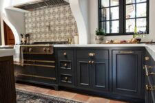 a Spanish style kitchen with a terracotta tile floor, navy cabinets, a large cooker and a built-in hood, dark-stained wooden beams