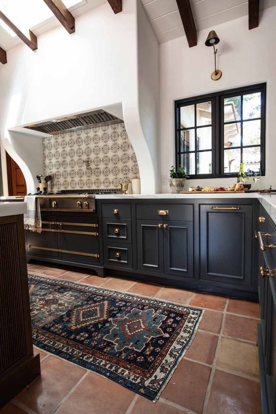 A Spanish style kitchen with a terracotta tile floor, navy cabinets, a large cooker and a built in hood, dark stained wooden beams