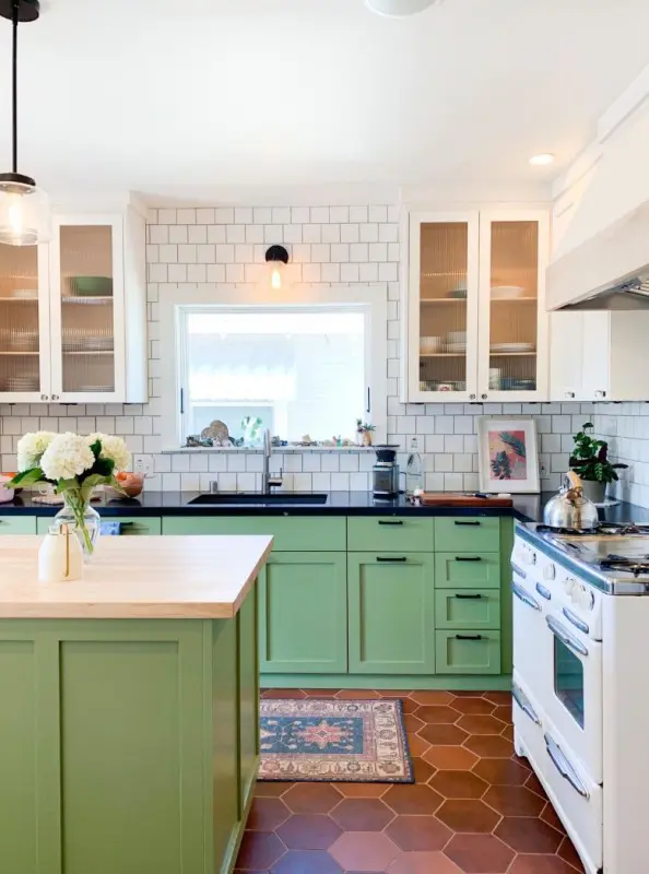 a bright modern kitchen with white fluted glass and green shaker style cabinets, black countertops, white square tile walls and a terracotta tile floor