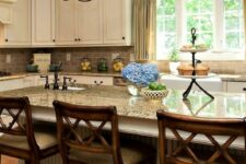 a creamy kitchen with shaker cabinetry, a taupe tile backsplash and beige granite countertops, vintage chandeliers and stained stools