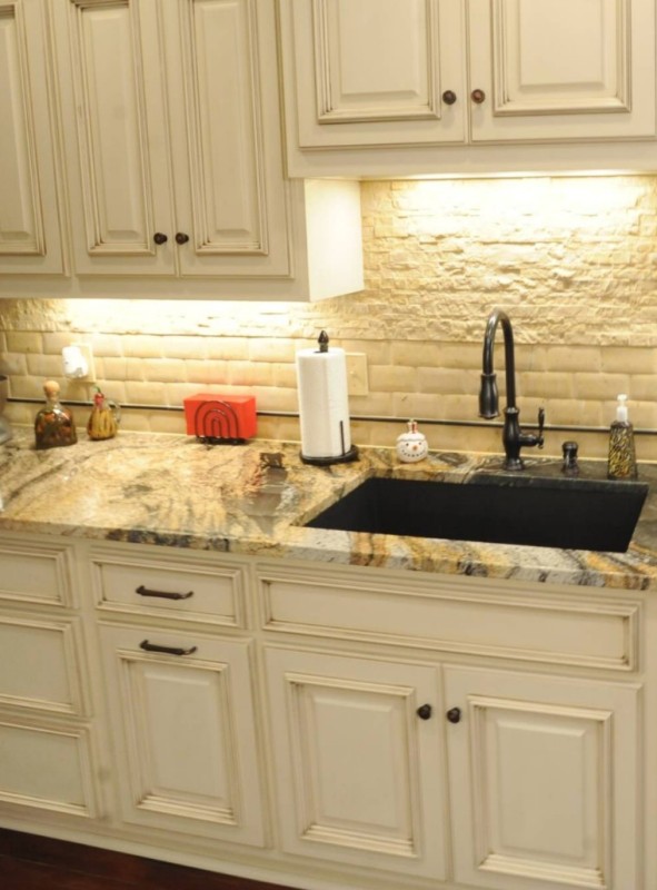 A creamy kitchen with shaker cabinets, a white tile and faux stone backsplash, built in lights and a gold granite countertop that makes a statement