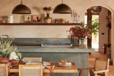a farmhouse kitchen with tan plaster walls, a terracotta floor, a concrete kitchen island, a wooden dining set and metal pendant lamps