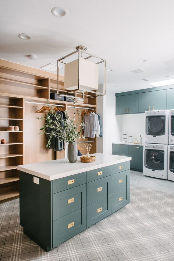 A large modern laundry mudroom with dark green cabinets, white countertops, a light stained storage unit, a chandelier and washing machines and dryers