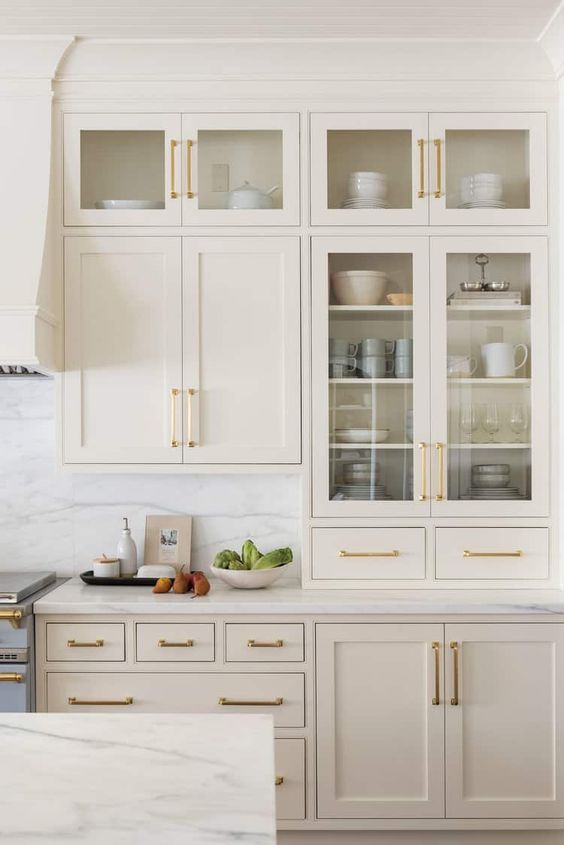 a lovely creamy kitchen with shaker and glass cabinets, white marble countertops and a backsplash and gold handles
