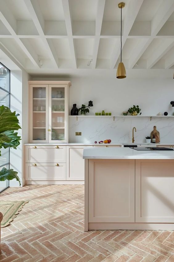 a modern farmhouse kitchen with a terracotta herringbone tile floor, blush cabinets, a white marble backsplash and pendant lamps