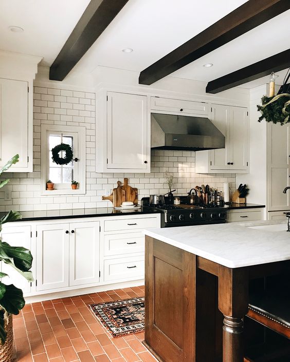 A modern farmhouse kitchen with a terracotta tile floor, a white subway tile backsplash, white shaker style cabinets, dark stained wooden beams