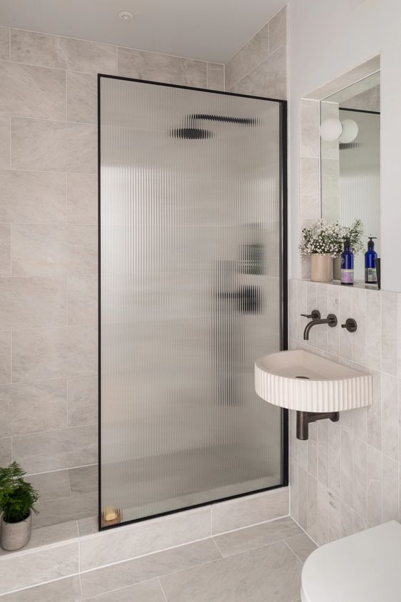 A neutral bathroom with stone tiles, a shower space with a fluted glass space divider, a wall mounted sink and a mirror with a shelf