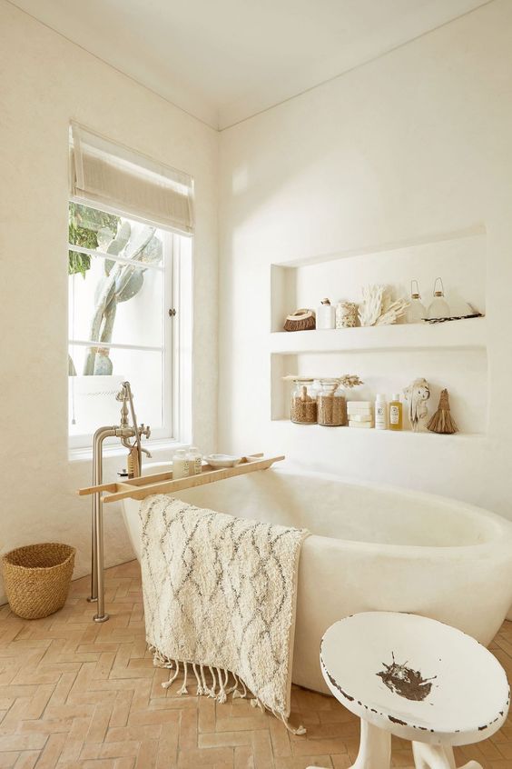 A neutral bathroom with white plaster walls with niches, a wabi sabi tub, a terracotta floor, a white stool and baskets