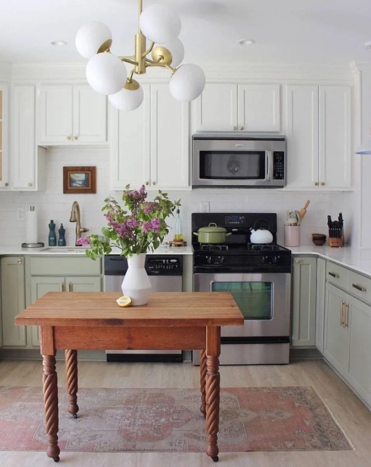 A two tone kitchen with white and sage green cabinets, a white tile backsplash and countertops and a small table as a kitchen island