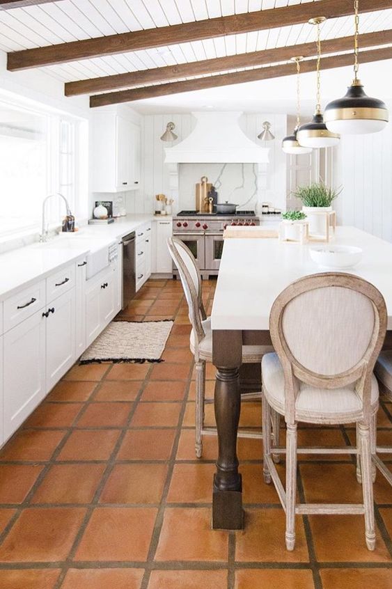 A vintage inspired white kitchen with only lower cabients, a large cooker and a hood, stained beams, a large table that is a kitchen island