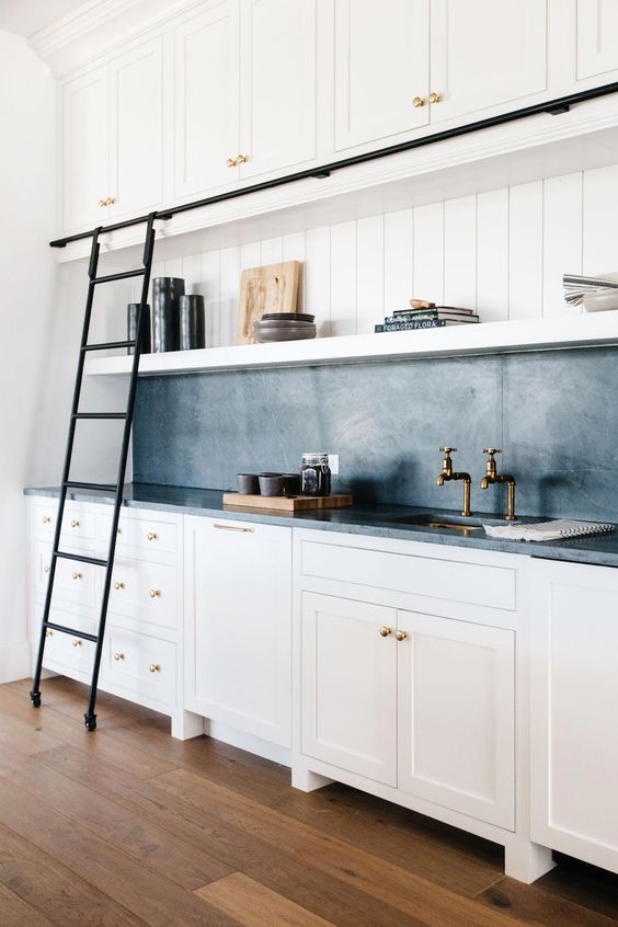 a white farmhouse kitchen with shaker cabinets, a black soapstone backsplash and countertops, open shelves and a black metal ladder