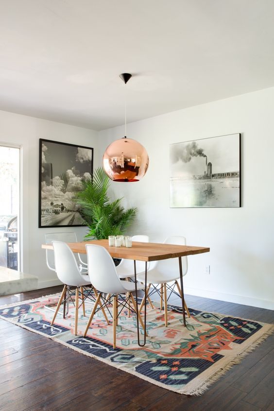 A mid century modern dining room with a hairpin leg dining table, white chairs, a printed rug, a copper pendant lamp and some art