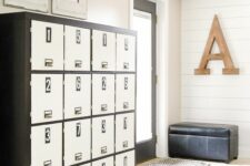 39 a neutral farmhouse mudroom with white beadboard walls, a large locker unit with numbers and a black leather upholstered bench