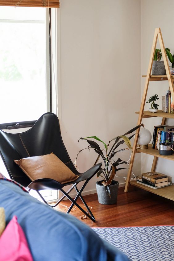 A black leather butterfly chair, a tan pillow, a ladder style bookshelf and a printed rug for a mid century modern space