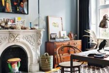 a boho eclectic home office with blue walls, an ornated French fireplace, printed layered rugs, a dark desk and a rattan chair, a stained sideboard
