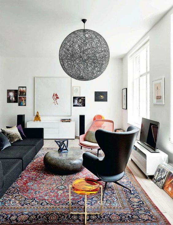 A boho living room with a low black sofa and a black chair, a stone like coffee table, a pink chair, a black yarn lamp