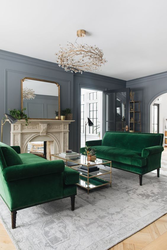 A catchy formal living room with a non working fireplace, green velvet sofas, a tiered glass coffee table and a unique chandelier