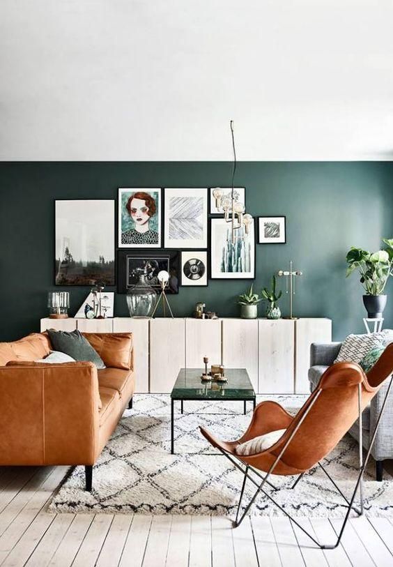 A chic mid century modern living room with a dark green accent wall, an amber leather sofa and a butterfly chair, a coffee table and a gallery wall
