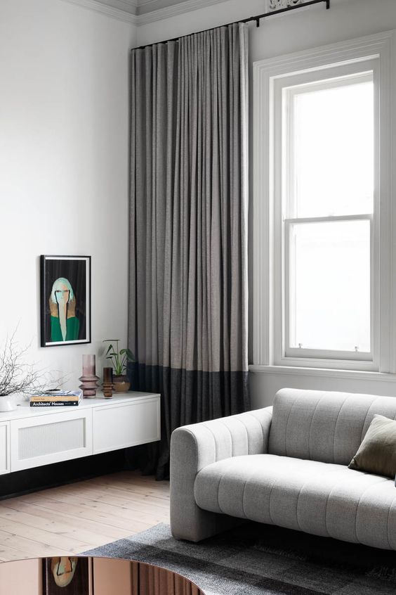 A contemporary living room with a double hung window, a grey sofa, a floating credenza, color block curtains and a printed rug