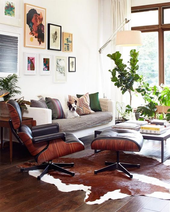 A cozy mid century modern living room with a neutral sofa with printed pillows, a large gallery wall, a black Eames lounger and ottoman and a coffee table