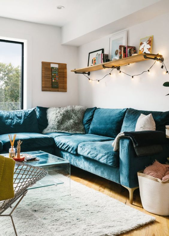 A cozy mid century modern living room with a navy velvet sectional, an acrylic coffee table, a metal chair, a shelf with lights and some pillows