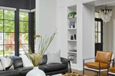 a double-height living room with a black leather sofa, amber chairs, a coffee table and some built-in shelves and greenery