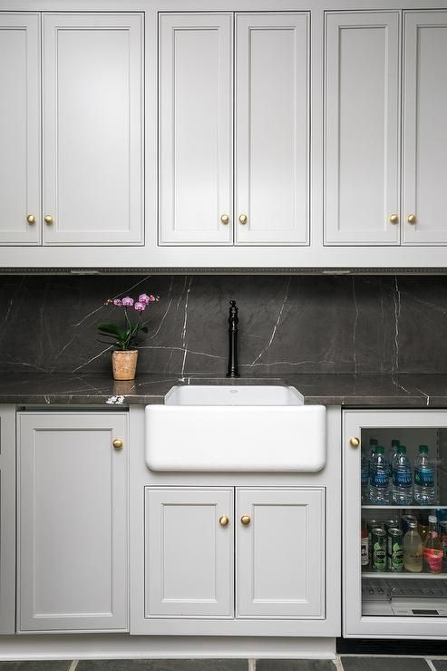 a dove grey kitchen with shaker cabinets, black soapstone countertops and a backsplash, gold knobs and a black faucet
