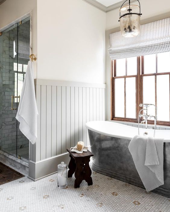 a farmhouse bathroom with paneling and penny tiles on the floor, a metal clad tub, stained double-hung windows, white towels