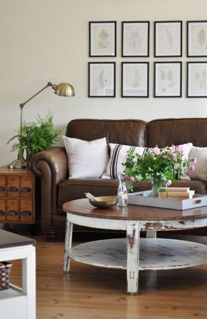 a farmhouse living room with a brown leather sofa, a shabby chic coffee table, a file cabinet, greenery and blooms and a gallery wall