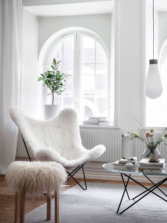 A light filled living room with arched windows, a white faux fur butterfly chair, a faux fur stool and a glass coffee table