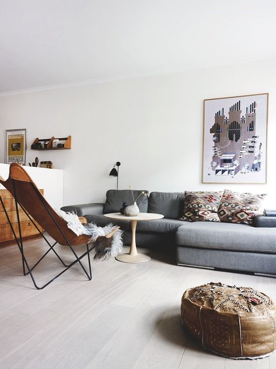 A mid century modern living room with a grey sectional, a brown leather butterfly chair, a leather pouf and printed pillows