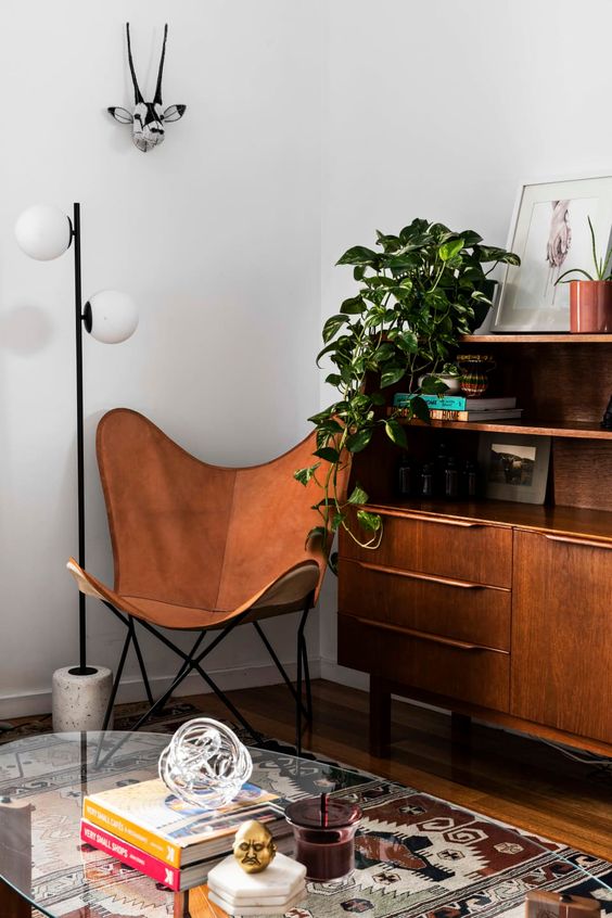 A mid century modern space with a stained sideboard, an amber leather butterfly chair, a printed rug, a glass coffee table and a plant