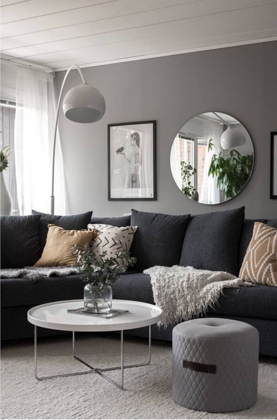 a monochromatic living room with a grey wall, a black sectional, printed pillows, artworks and a round mirror, a coffee table and a pouf
