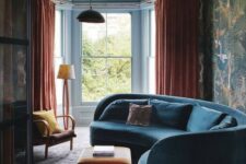 a moody and bold living room with a curved blue velvet sofa, orange textiles and moody bird print wallpaper on one wall