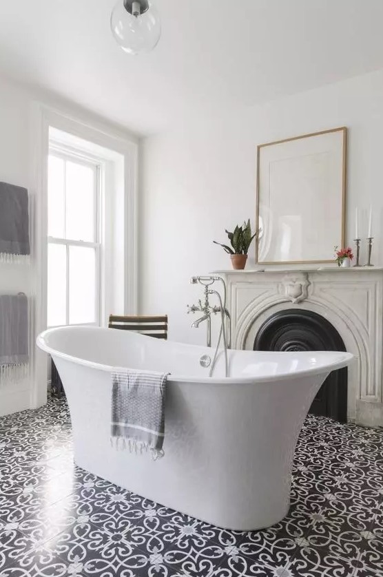 A refined and chic bathroom with a vintage French fireplace with a stone mantel, a free standing tub, some art and beautiful tiles on the floor