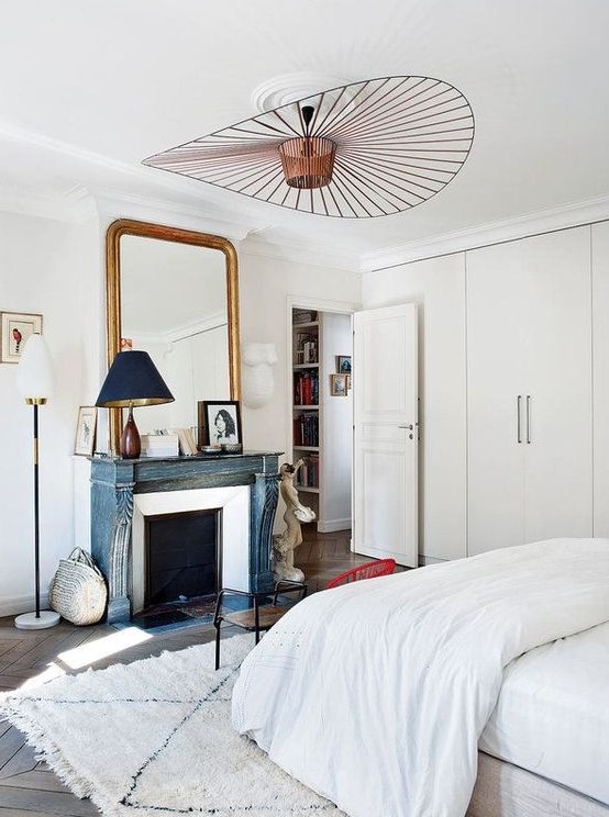 A serene Parisian bedroom with built in wardrobes, a French fireplace with a blue mantel, a large mirror in a gilded frame and a bed with neutral bedding